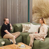 Grøn udendørs hjørnesofa med bløde puder, hvor to personer slapper af ved et træbord med drinks og snacks omgivet af klitter og marehalm.