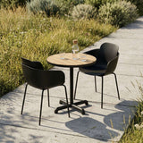 sackit Kirra cafésæt med rundt udendørs spisebord i træ og to sorte stole på en solrig terrasse omgivet af grønne planter
