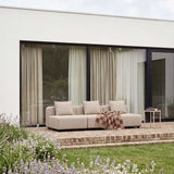 Beige trepersoners loungesofa fra sackit på en terrasse med puder og ternet tæppe foran et moderne hus og en blomstrende have