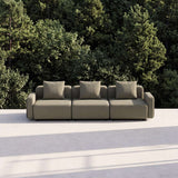 Trepersoners udendørs loungesofa fra sackit med tre hynder i neutral beige farve omgivet af frodige grønne træer på en solrig terrasse