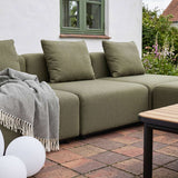 Olivengrøn modulsofa med bløde hynder og ternet plaid på en hyggelig terrasse omgivet af grønne planter og et lavt havebord