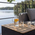 Udendørs sofabord med lamelbordplade i teak foran en grå loungesofa med kande og glas ved en rolig sø fra sackit