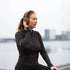 Kvinde i sort træningstøj med brune over ear høretelefoner fra sackit ved vandet og med hånden på ørekoppen til løb og aktiv brug