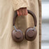 Brune over-ear høretelefoner fra sackit holdes i hånden foran en beige frakke. Det polstrede design og de runde ørepuder fremhæver komfort og stil.