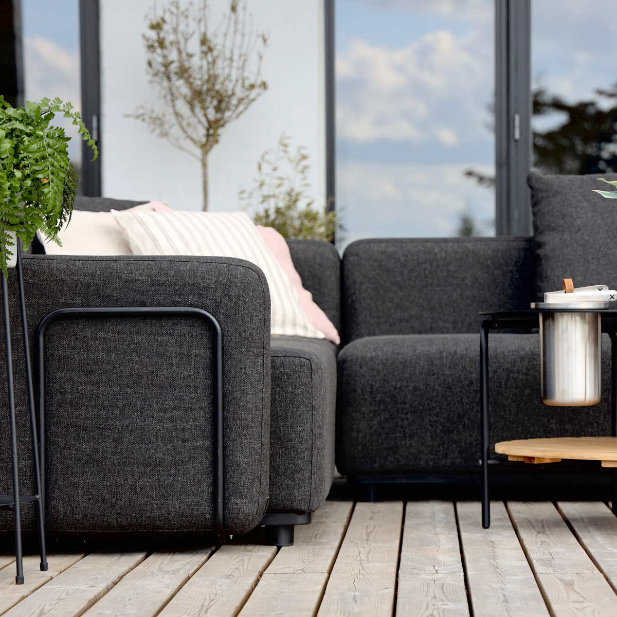 Mørkegrå modulsofa med armstøtte og bløde puder på en træterrasse med plante og lille sidebord i et lyst nordisk uderum