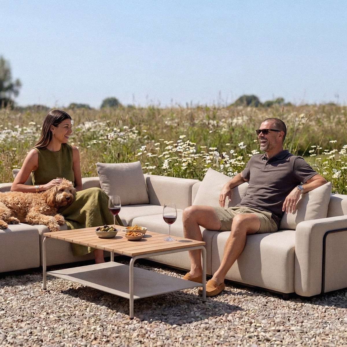 Udendørs hjørnesofa med lyse hynder hvor et par slapper af med hund i blomstrende natur omkring et lavt træbord datingside