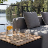 Grå sackit Cobana loungesofa med teakbord og glas på en solrig terrasse ved søen. Dansk design til afslappende udendørsliv