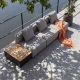 Grå modulopbygget loungesofa fra sackit på terrasse med sidebord i teaklook puder orange tæppe og glas i skyggen af grønne træer