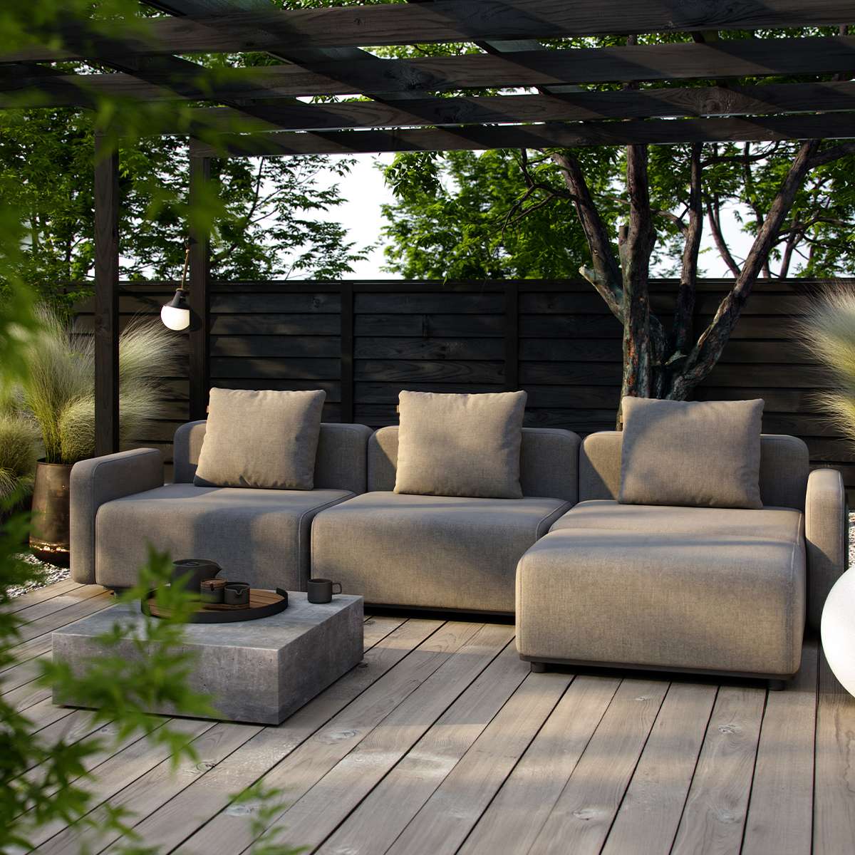 Modulopbygget Cobana loungesofa i gråt tekstil med puder på træterrasse under pergola omgivet af grønne planter i en rolig have