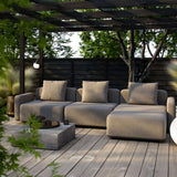 Modulopbygget Cobana loungesofa i gråt tekstil med puder på træterrasse under pergola omgivet af grønne planter i en rolig have