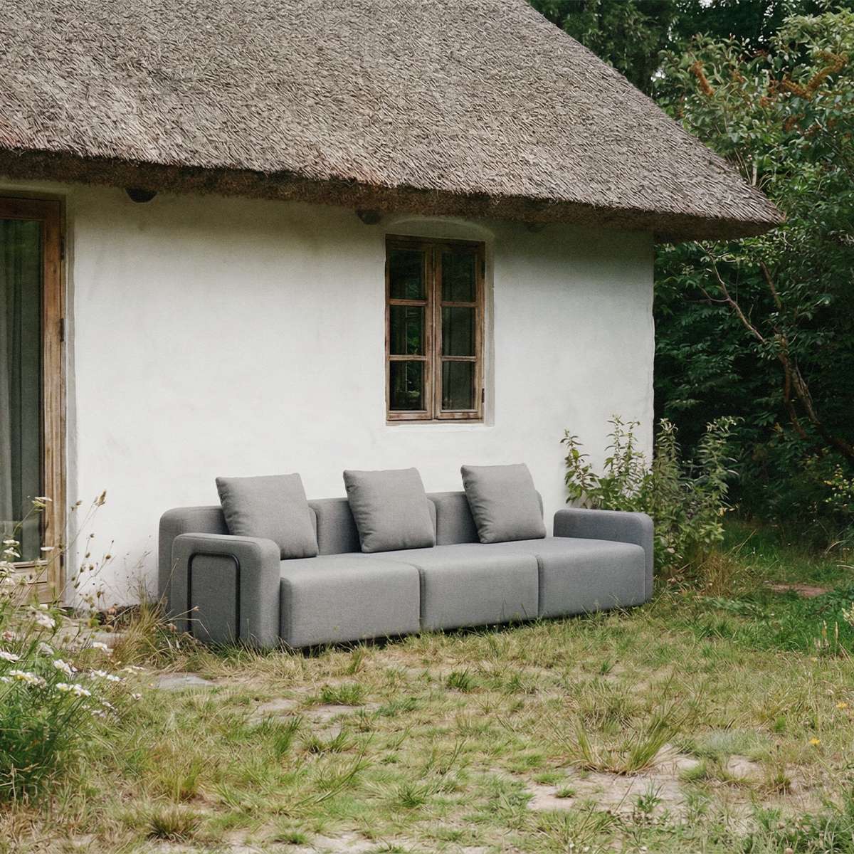 Grå trepersoners udendørs sofa fra sackit placeret foran et stråtækt hus i en frodig have med bløde puder og skandinavisk design
