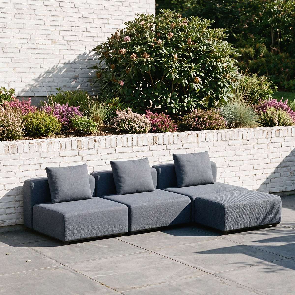 Mørkegrå modulopbygget udendørs loungesofa med puf og puder på en solrig terrasse ved en hvid mur designet i Danmark af sackit