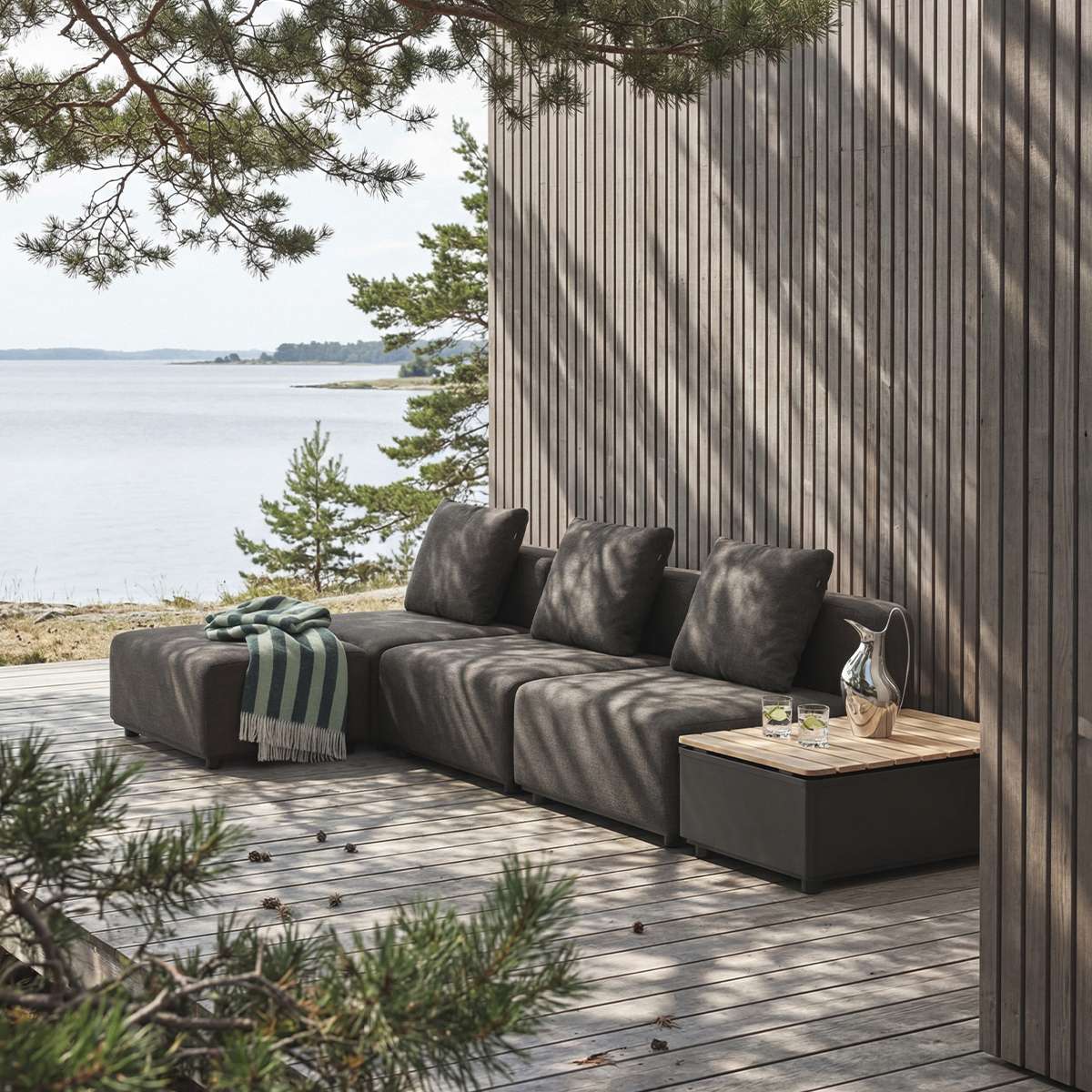 sackit modulopbygget udendørs loungesofa med puf i mørkt tekstil på træterrasse ved vandet med puder og sidebord