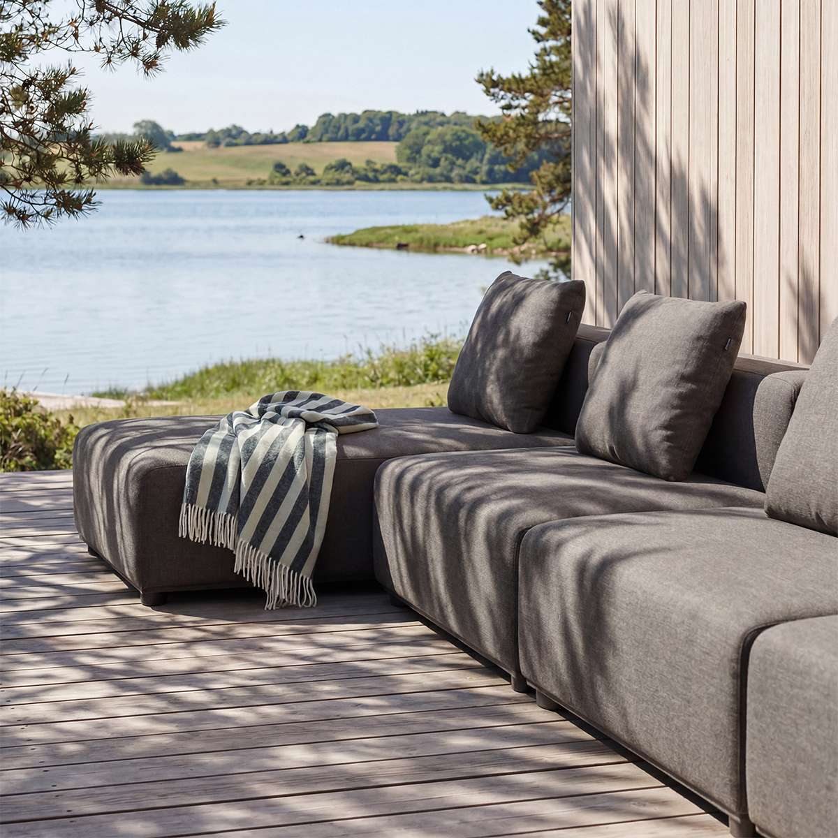 Grå modulopbygget loungesofa med chaiselong på træterrasse ved søen med bløde puder og stribet tæppe fra sackit