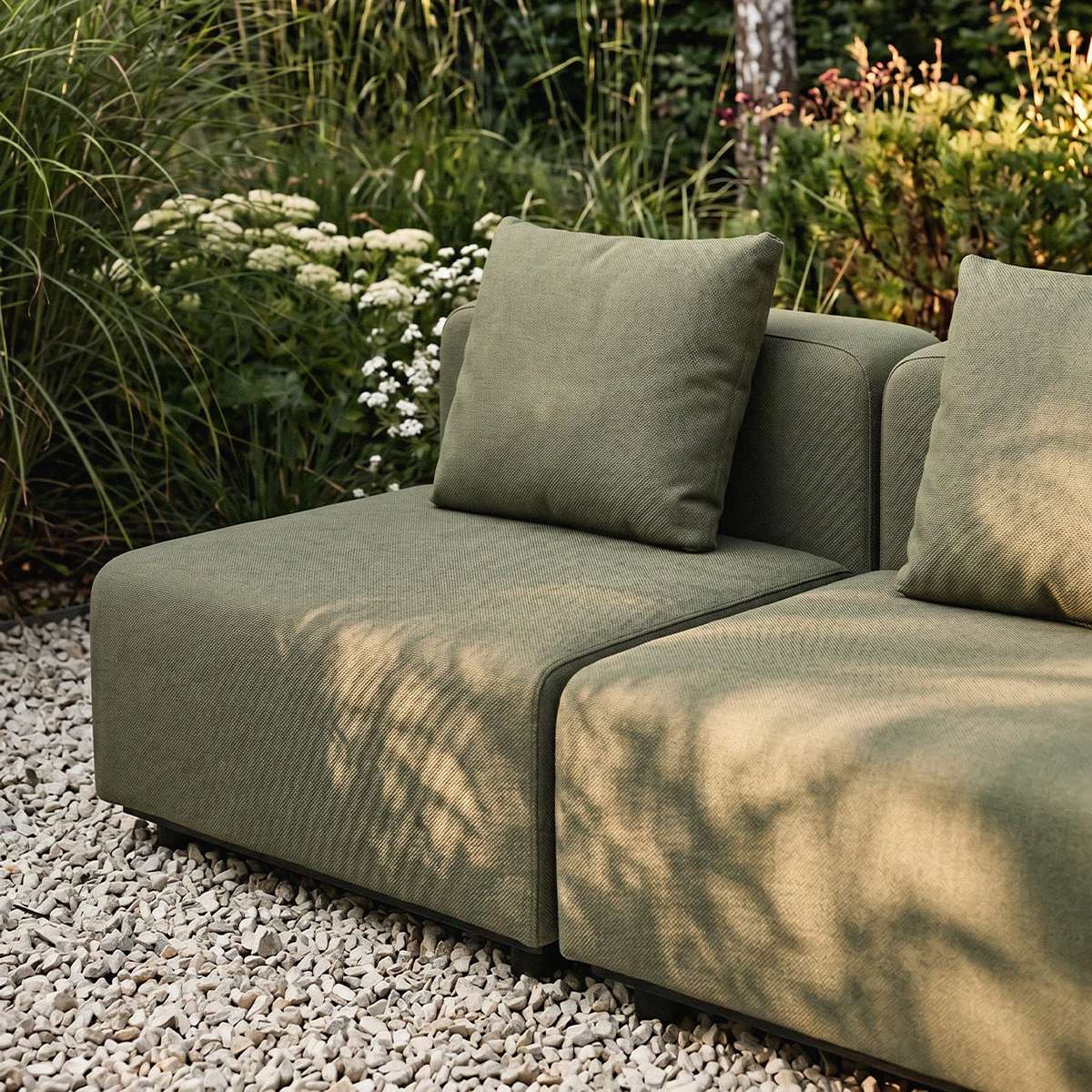 Grøn modulopbygget hjørnesofa fra sackit på en grusbelagt terrasse omgivet af frodige planter og bløde puder i nordisk design