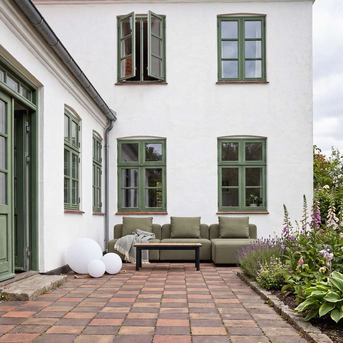Olivegrøn Cobana loungesofa med tre siddepladser og sofabord på en hyggelig gårdhave omgivet af grønne planter og blomster
