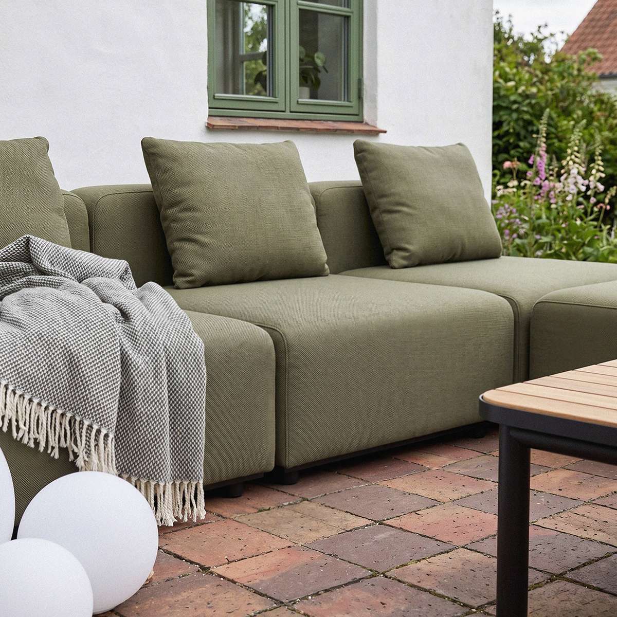 Olivengrøn modulsofa med bløde hynder og ternet plaid på en hyggelig terrasse omgivet af grønne planter og et lavt havebord
