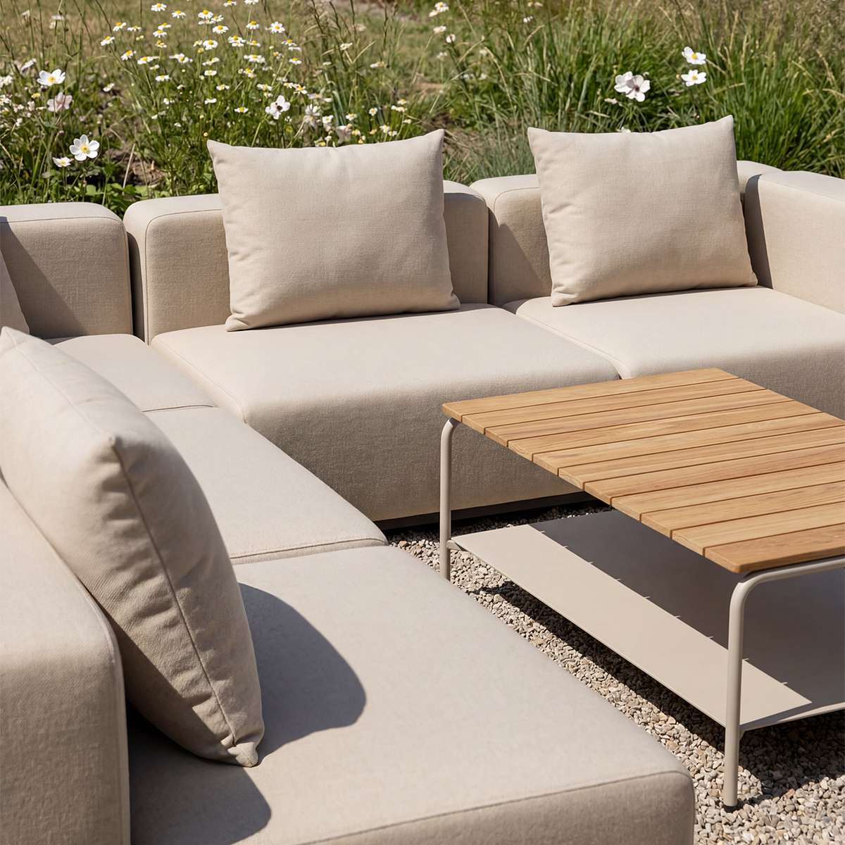 Beige modulopbygget udendørs hjørnesofa med bløde puder og matchende træbord på en solrig terrasse omgivet af blomstrende græs