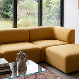 Sennepsgul Fjord sofa med matchende puf i en lys stue foran store vinduer og et glasbord med dekorative genstande fra sackit