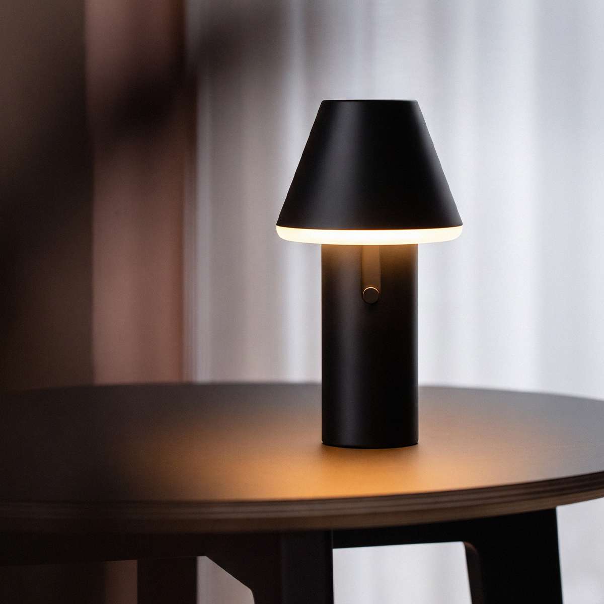 Sort trådløs bordlampe fra sackit med konisk skærm og blødt varmt lys på et mørkt træbord i elegant nordisk interiør