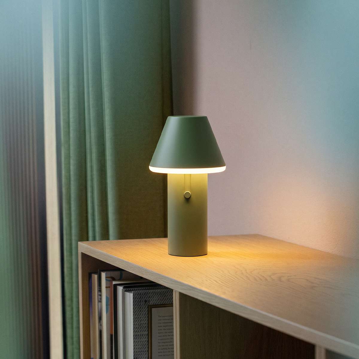 Grøn trådløs bordlampe fra sackit på en træhylde med bøger og gardin i baggrunden og varmt lys der skaber en hyggelig stemning