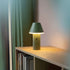 Grøn trådløs bordlampe fra sackit på en træhylde med bøger og gardin i baggrunden og varmt lys der skaber en hyggelig stemning
