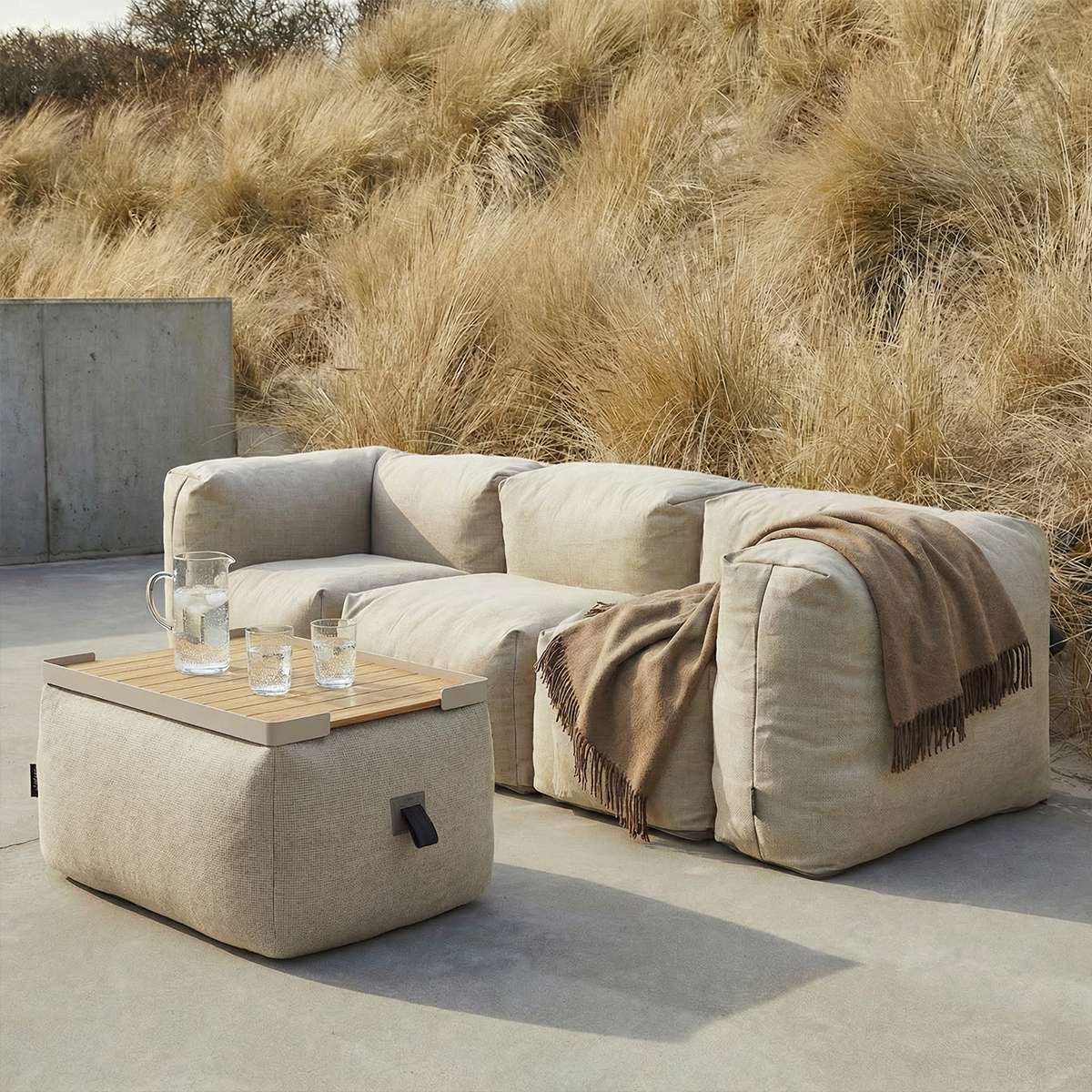 Beige udendørs pouf fra sackit med træbakke foran en blød loungesofa på en terrasse omgivet af marehalm