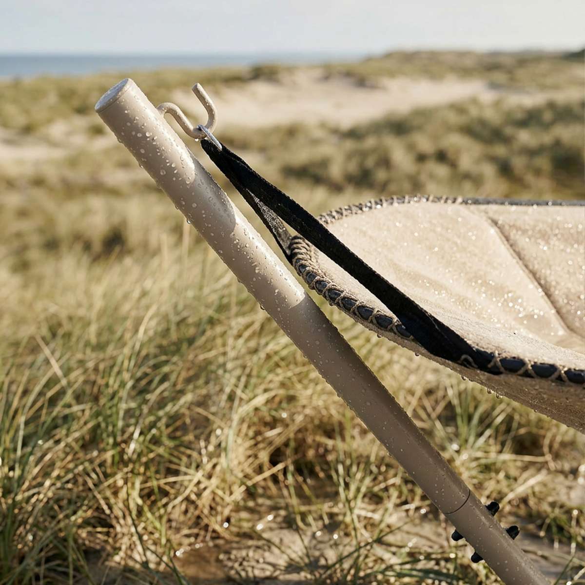 Detalje af beige Cobana hængekøje med stativ og sorte ophængsbånd i klitlandskab ved havet, med vanddråber på stoffet