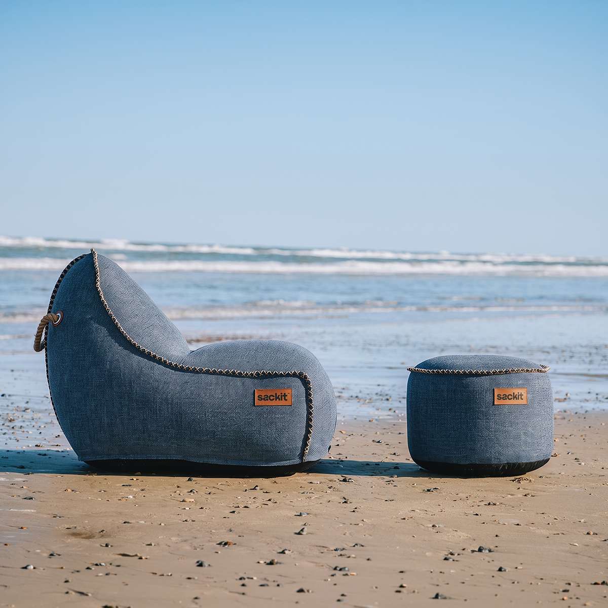 sackit Cobana loungestol og puf i blågråt stof på en strand ved havet under en klar blå himmel