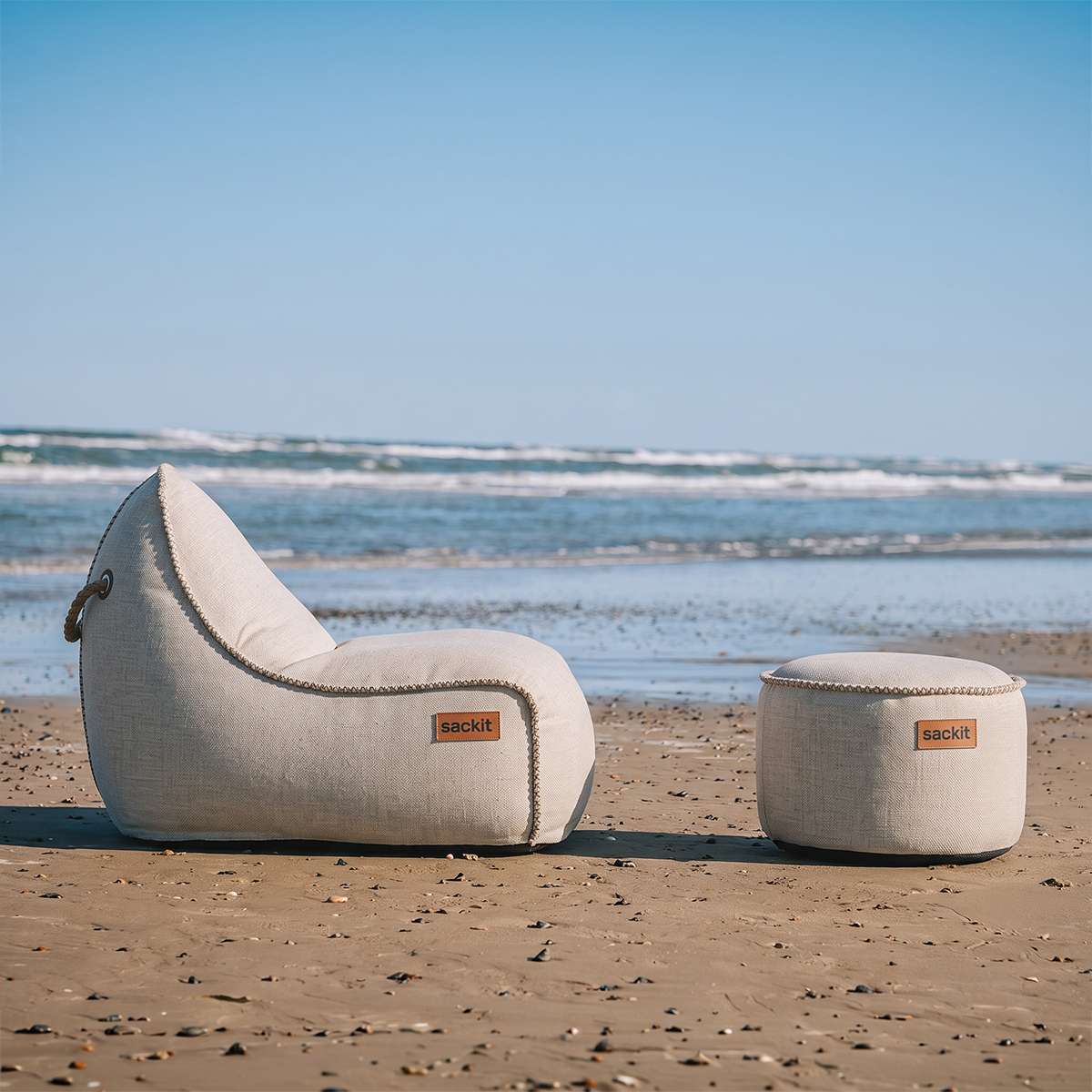 sackit Cobana loungestol og puf i beige står på en sandstrand med havet i baggrunden som stilrent udendørs loungesæt til nordisk vejr