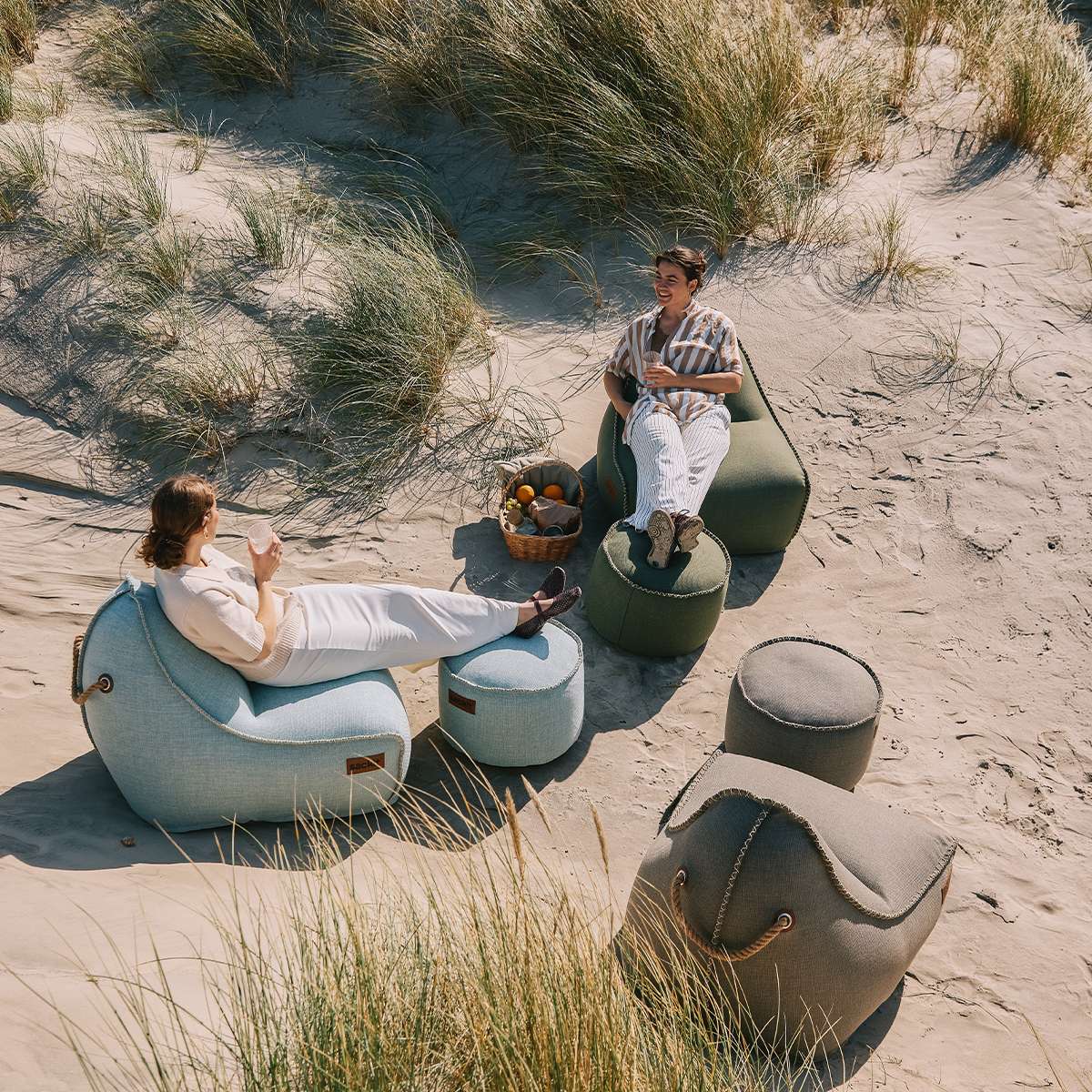 To personer slapper af i bløde Cobana sækkestole og puffer på en solrig sandstrand omgivet af marehalm fra sackit
