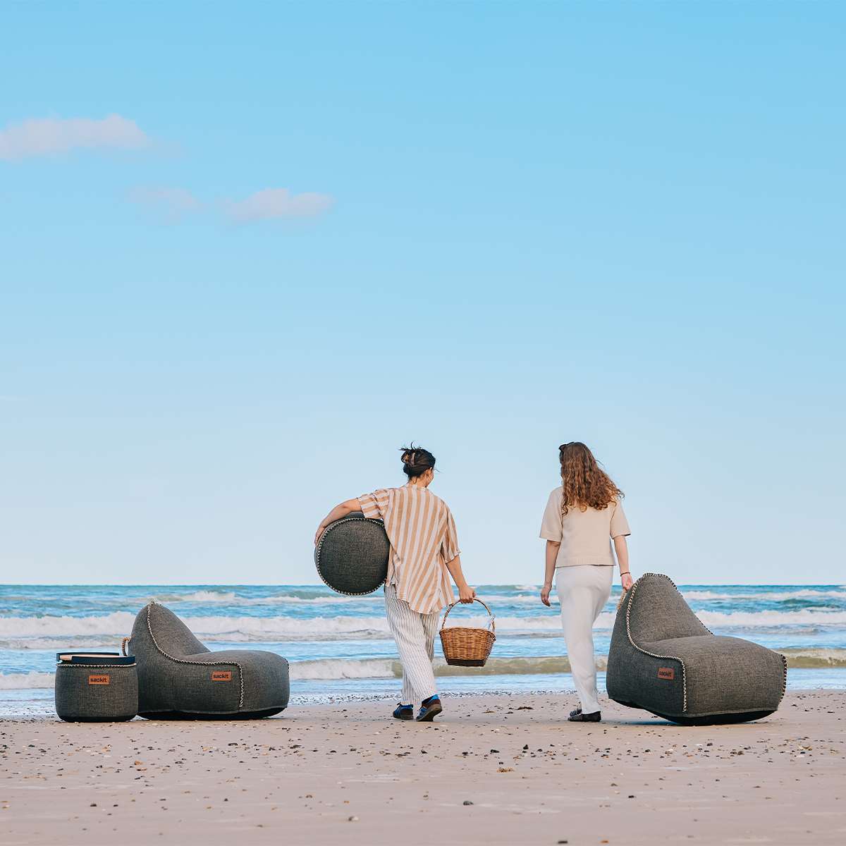 To mørke Cobana loungestole og puffer fra sackit på stranden med havet i baggrunden og et par på vej langs vandkanten