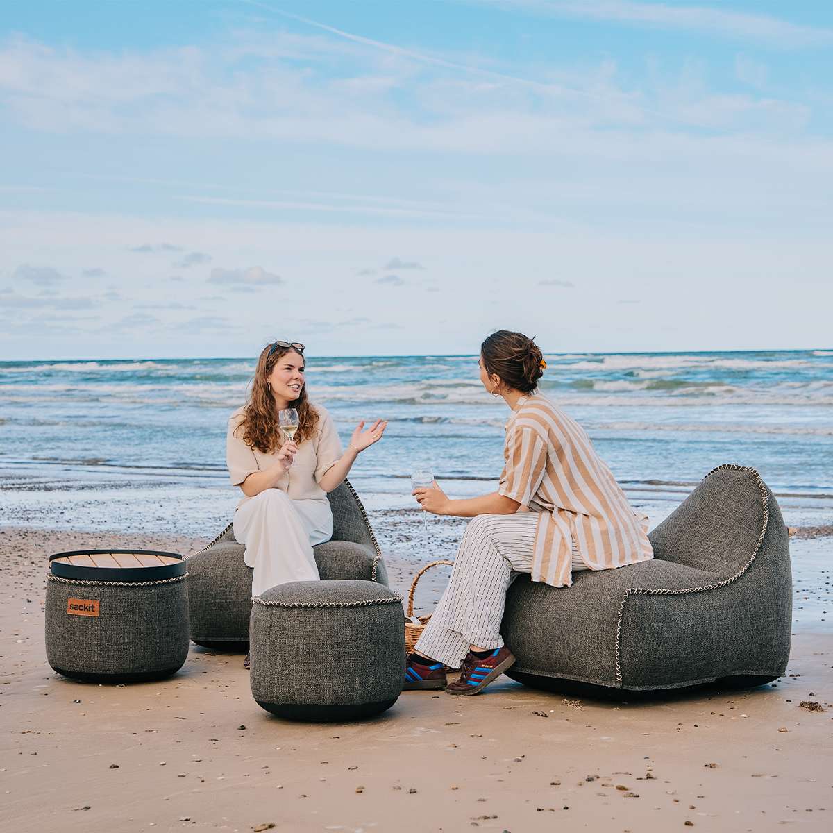 To kvinder nyder udsigten ved havet i en sackit loungestol og to grå puffer på stranden i naturlige og afslappede omgivelser