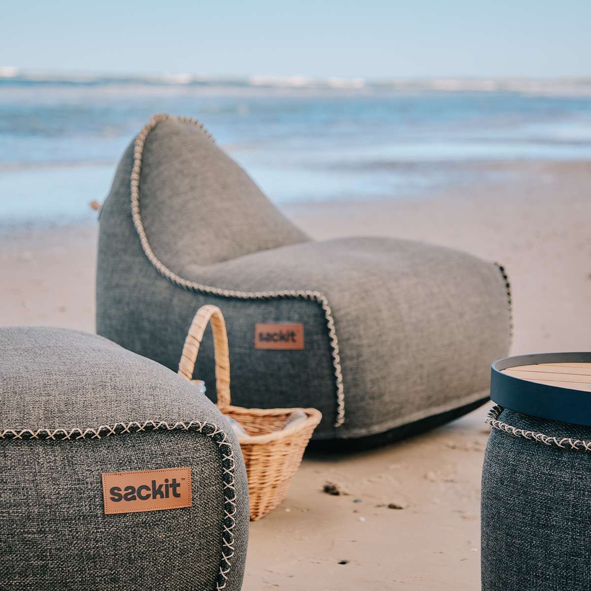 Grå sackit Cobana loungesækkestol og puf på stranden ved havet med flettede detaljer en kurv og et rundt sidebord