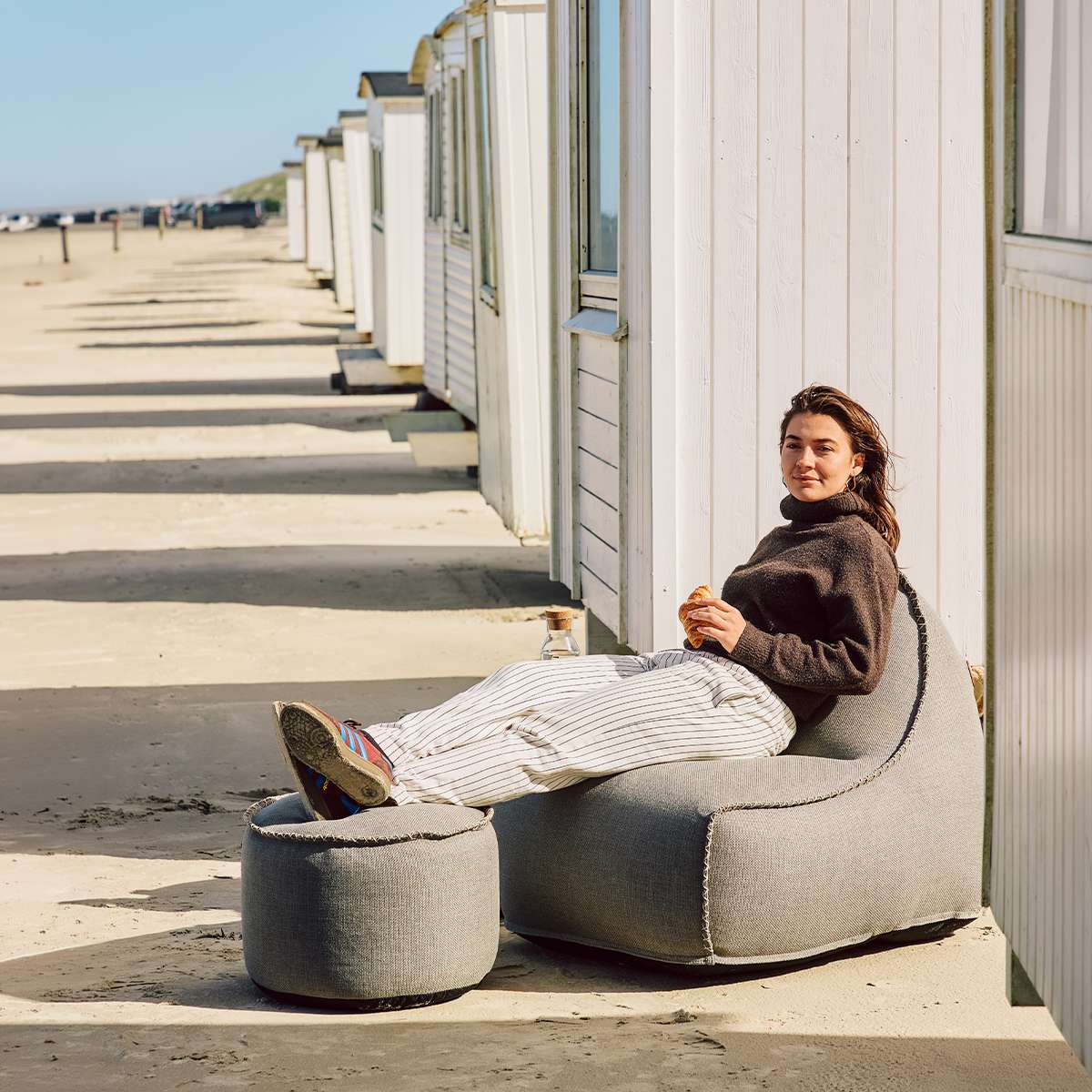 Kvinde slapper af i en grå udendørs sækkestol fra sackit med matchende puf foran hvide strandhytter på en solrig strand.