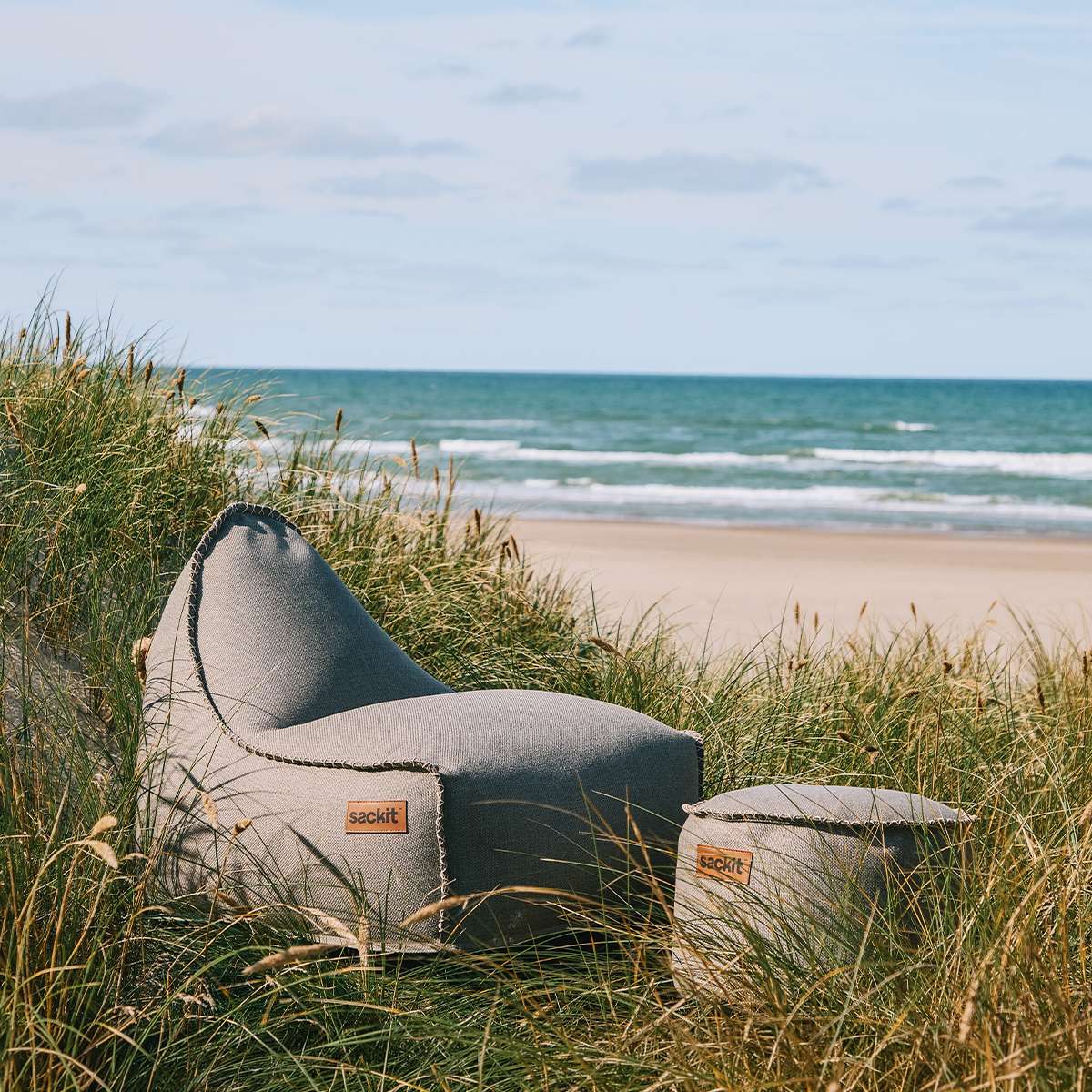sackit Cobana loungestol og puf i gråt stof placeret mellem marehalm ved stranden med havet i baggrunden, velegnet til nordisk udendørs afslapning