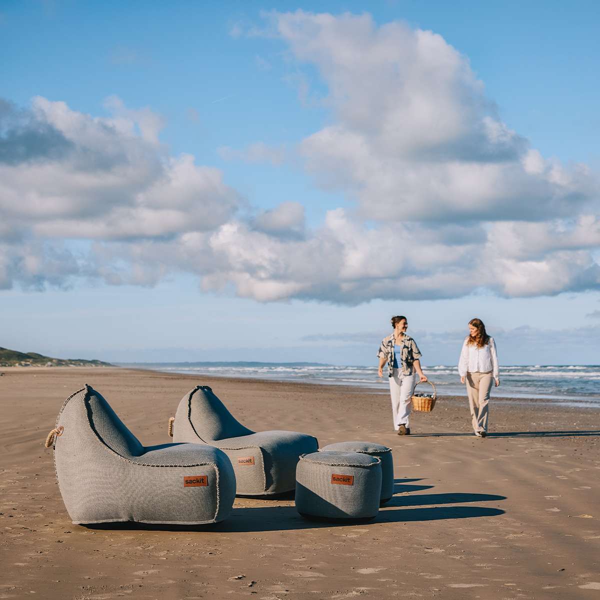 To grå sackit Cobana loungestole og en matchende puf står på stranden med havet i baggrunden og to personer på vej mod møblerne