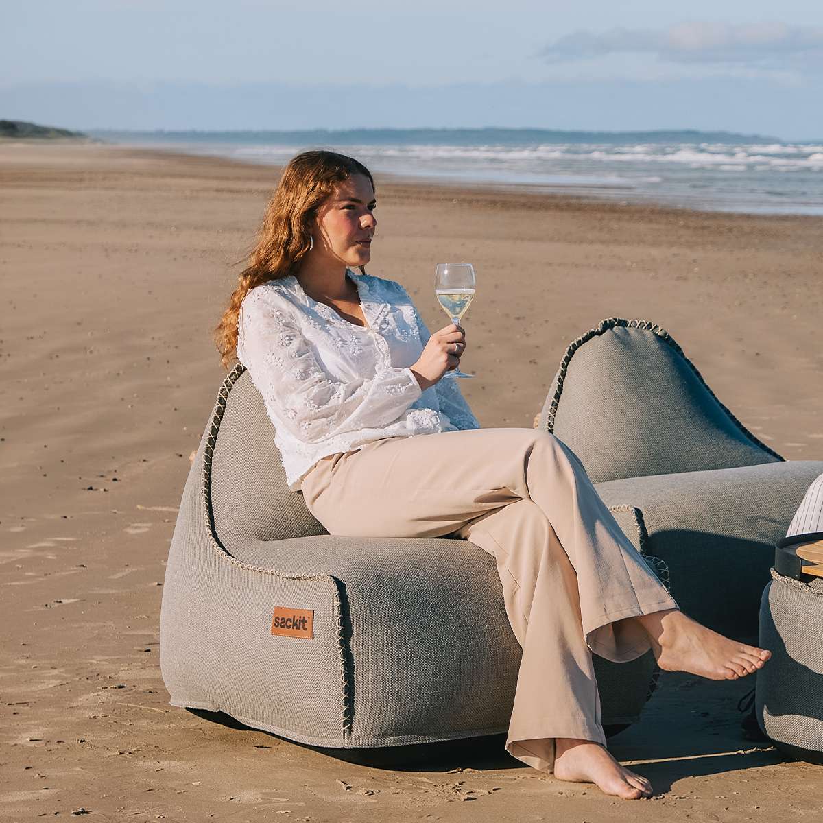 sackit udendørs loungestol og puf på stranden med kvinde der nyder et glas vin i det vejrbestandige design til nordisk vejr