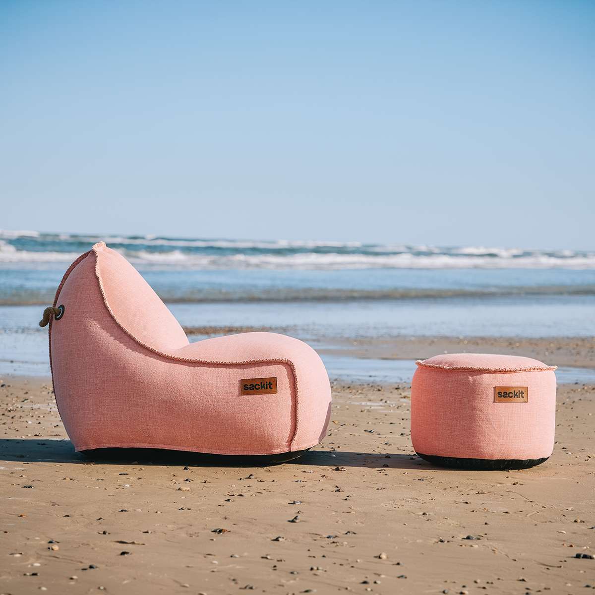 Rosa sackit lounge stol og puf placeret på en sandstrand med hav og blå himmel i baggrunden. Scandinavian design til afslappende udendørs ophold.