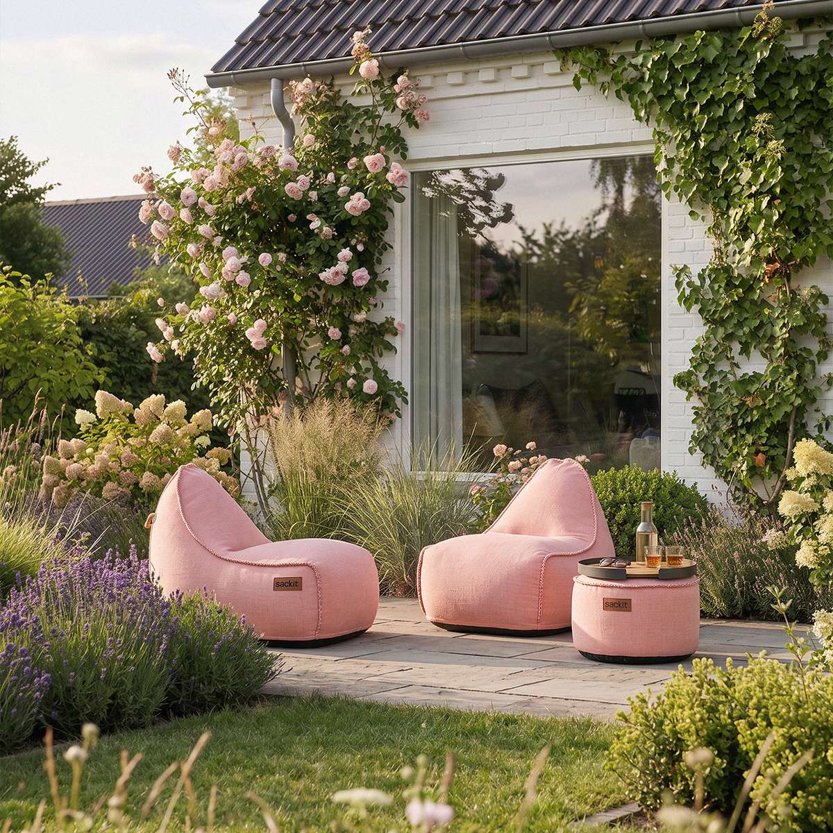 To rosa sackit loungestole med matchende pouf på en terrasse omgivet af blomstrende planter og frodigt grønt i en hyggelig have