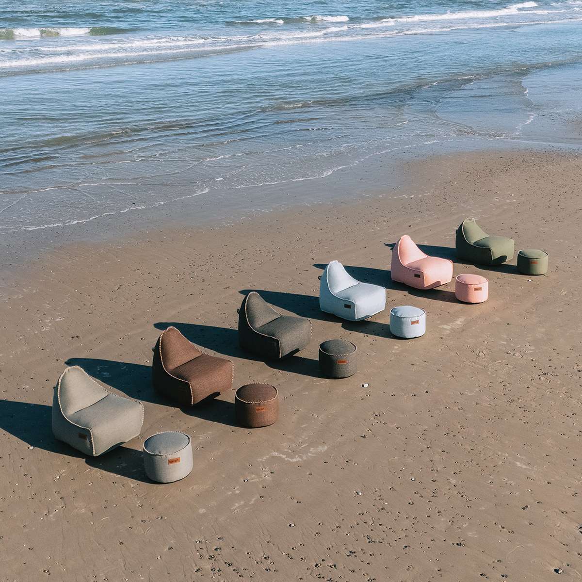 Flere farverige udendørs sækkestole med matchende puffer fra sackit står på stranden ved havet og skaber en afslappet loungeoplevelse i det fri
