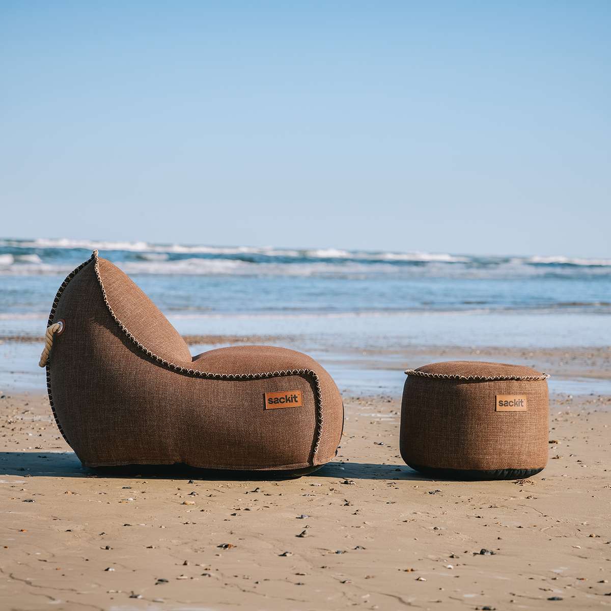 sackit Cobana loungestol med matchende puf i brunt stof på en solrig strand med havet i baggrunden til afslappet udendørs loungekomfort