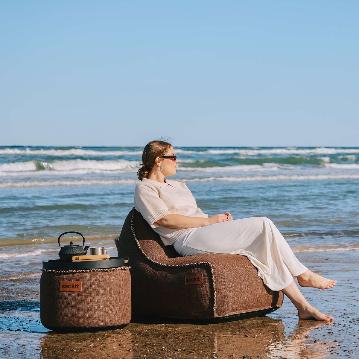 sackit Cobana loungestol og puf i brun tekstil ved stranden med en kvinde der nyder udsigten over havet